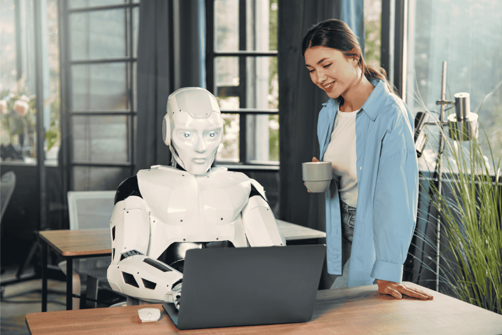 A robot sits working at a laptop while a woman drinks a hot drink. 