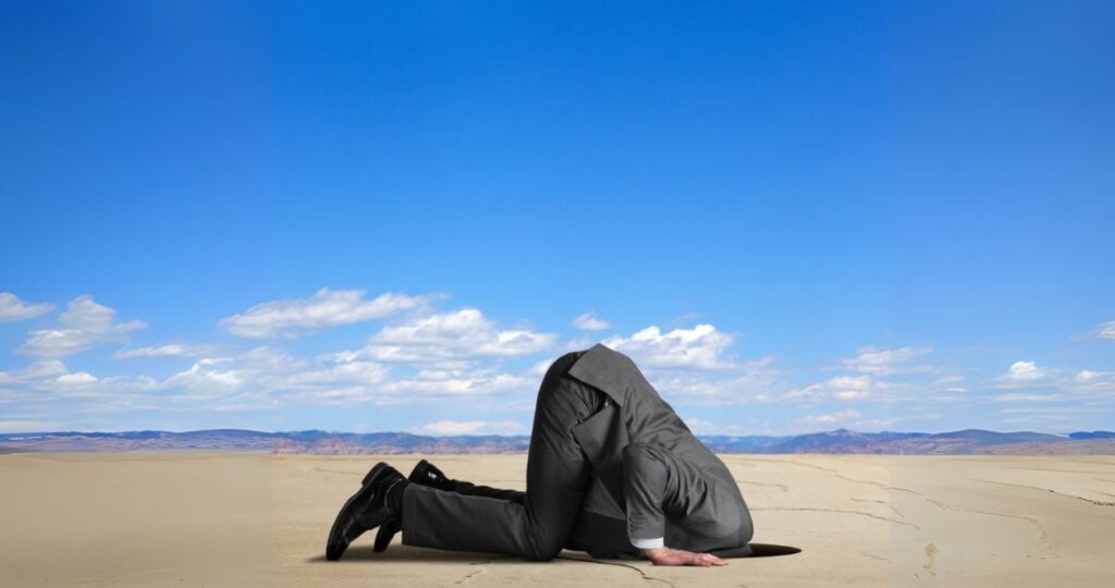 A man in a suit with his head buried in the sand, with a clear blue sky overhead